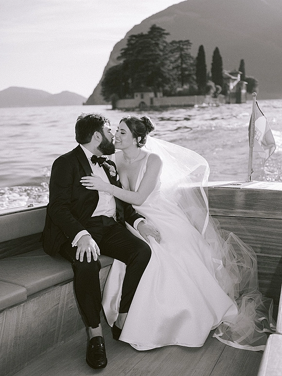 Wedding kiss portrait of bride and groom on boat, her long veil flowing as they lean in above a mountain lake with islands behind