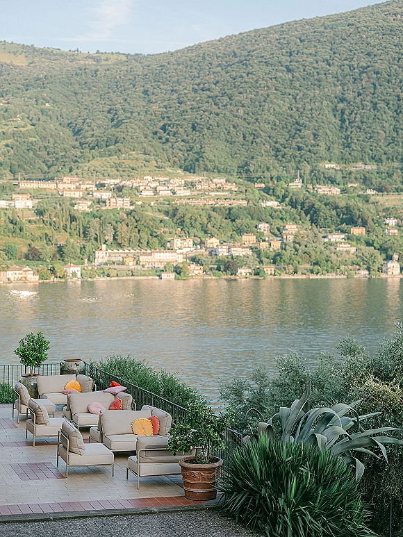 Outdoor lounge seating with sofas and colorful throw pillows on a tiled terrace, with terracotta planters and lake-and-mountain views beyond