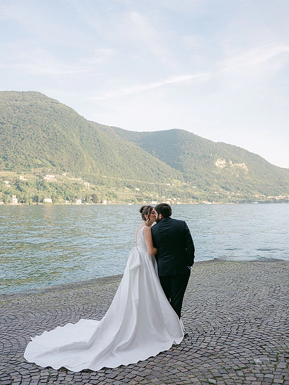 Wedding kiss portrait of bride and groom kissing, her long train flowing on cobblestones by a mountain lake promenade under open sky