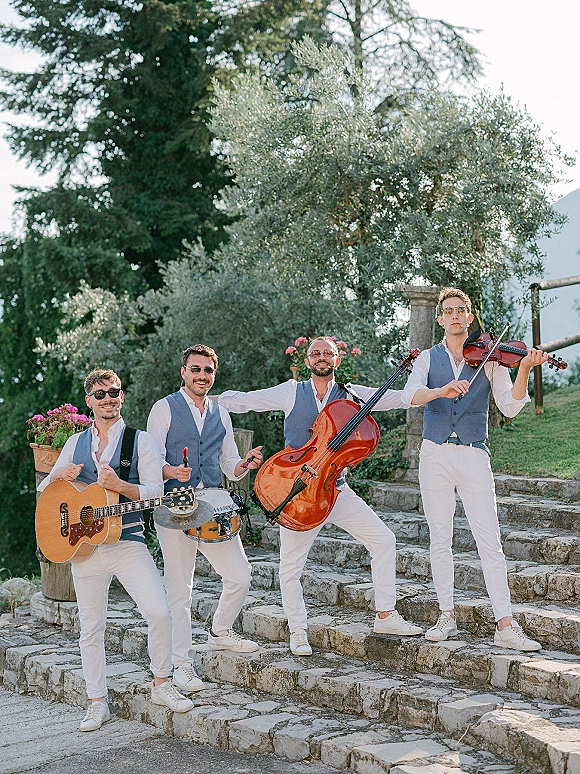 Wedding band in matching vests and sunglasses posing with guitar, cello, violin and snare drum on stone steps by greenery outdoors