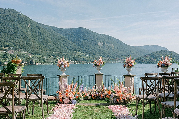 Ceremony setup with floral arrangements lining a rose petal aisle on a terrace, crossback chairs facing a lake and mountains view