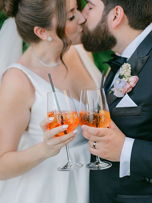 Wedding kiss portrait of bride and groom kissing while toasting cocktails with straws, veil and black tuxedo details against greenery backdrop
