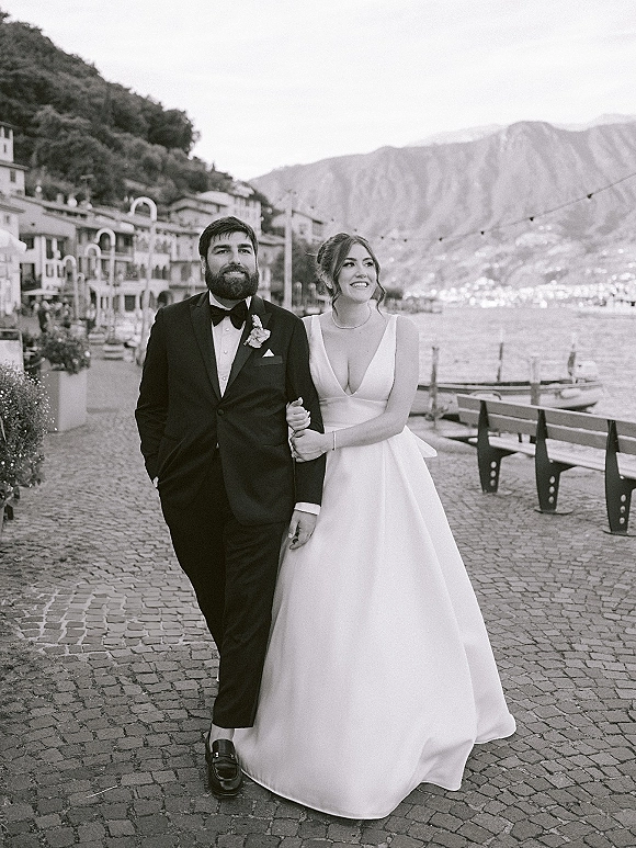 Couple portrait in a black and white wedding portrait style, bride in deep V dress holding groom’s arm on a lakeside promenade with mountains and boats behind