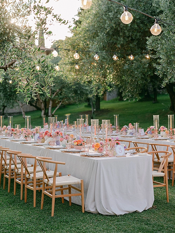 Reception tablescape for an outdoor wedding reception with long white-linen banquet tables, wooden chairs, candles, florals, and string lights on a garden lawn