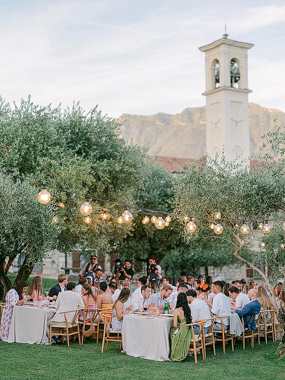 Outdoor wedding reception with round linen tables under string lights, taper candles and florals beneath olive trees with mountain views
