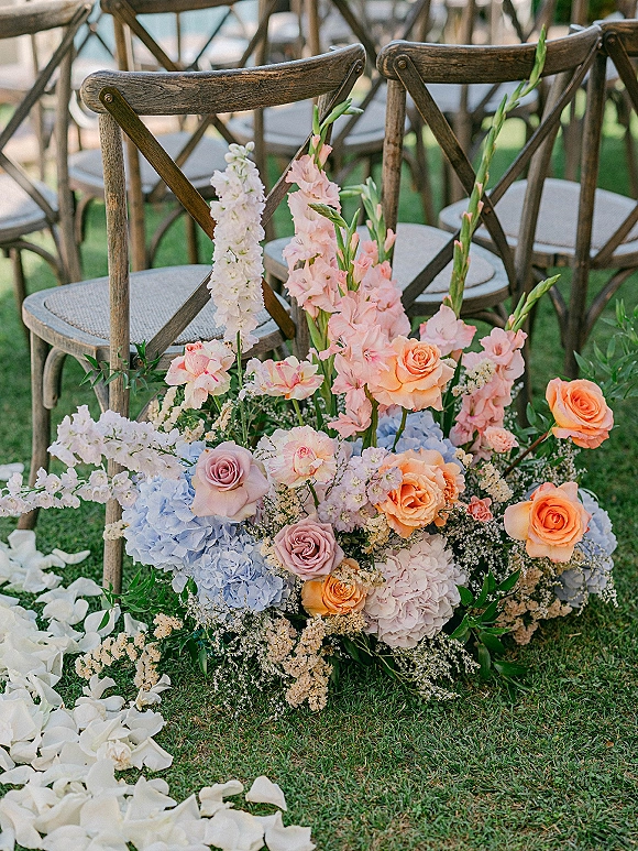 Ceremony aisle flowers in a low cluster of roses, hydrangea, gladiolus and greenery by wood cross-back chairs on a grass lawn