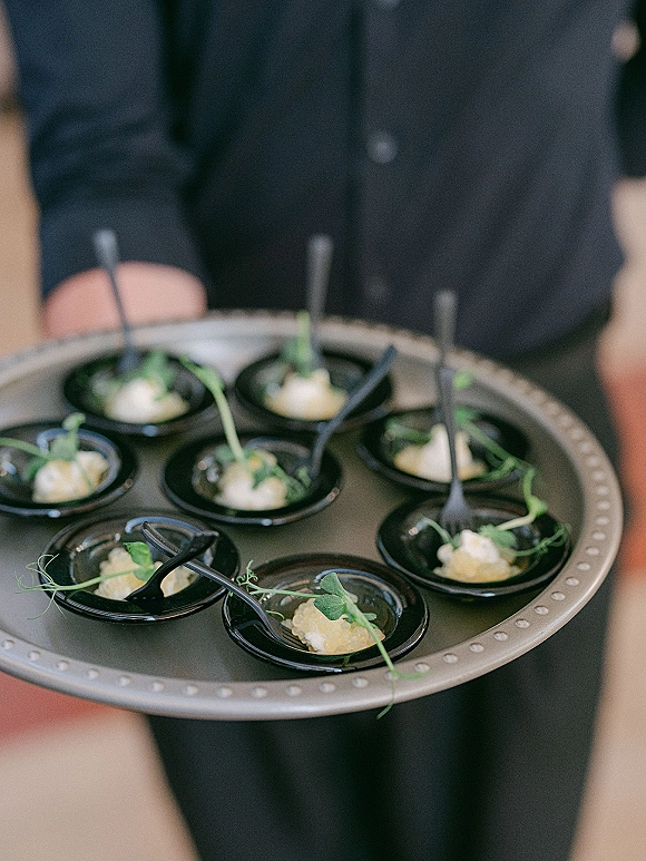 Wedding cocktail hour appetizers on a passed appetizer tray with black tasting spoons, microgreens garnish, and small bowls in a blurred indoor setting