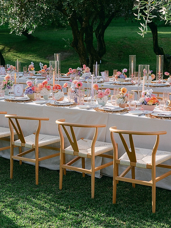 Reception tablescape with pastel florals in glass cylinder vases, taper candles, blush glassware, and rattan chargers on a garden lawn