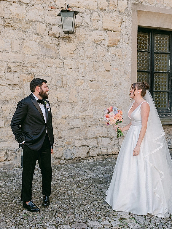 Couple portrait of bride and groom smiling, bride holding bouquet with veil and satin dress beside a stone wall and lantern outdoors