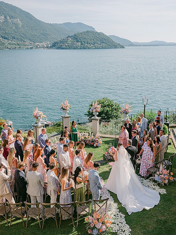 Wedding ceremony as the bride walks down the aisle toward the groom, her long veil trailing over a white petal runner by a mountain lake terrace
