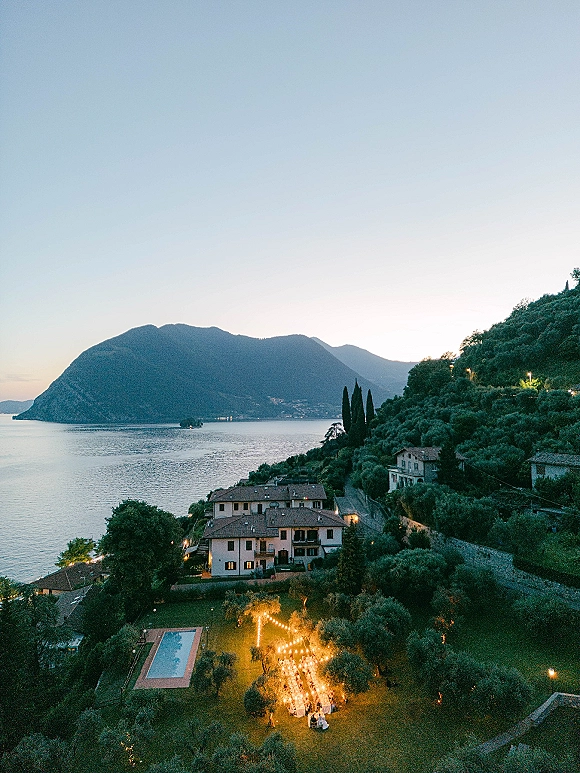 Outdoor reception setup with string lights over long banquet tables, set on a villa lawn by a pool with lake and mountains at dusk