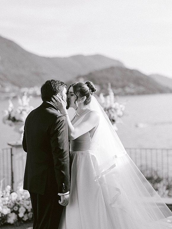 Wedding kiss portrait of bride and groom kissing, long veil blowing as she cups his face on a terrace with mountains and lake behind