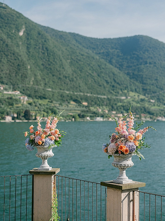 Wedding ceremony florals in stone urns with roses, hydrangeas, and gladiolus lining a terrace railing overlooking a lake and mountains
