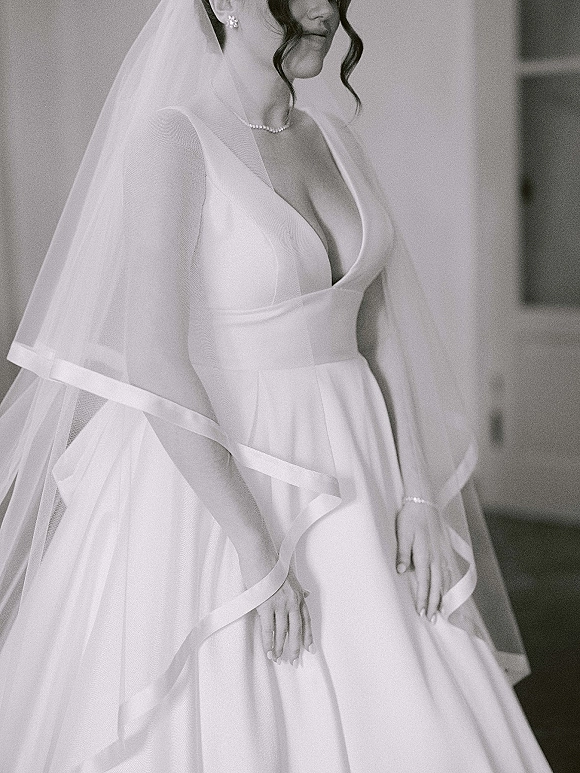 Bridal portrait in black and white of a bride in a deep V wedding dress, veil over shoulders, wearing a pearl necklace by a doorway