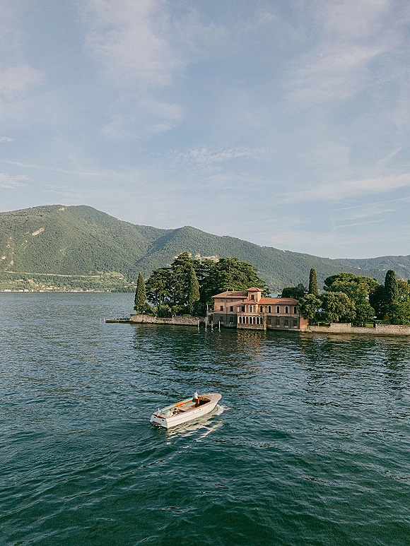 Wedding boat ride with bride and groom on a wood speedboat, gliding past a lakefront villa with mountains and trees behind