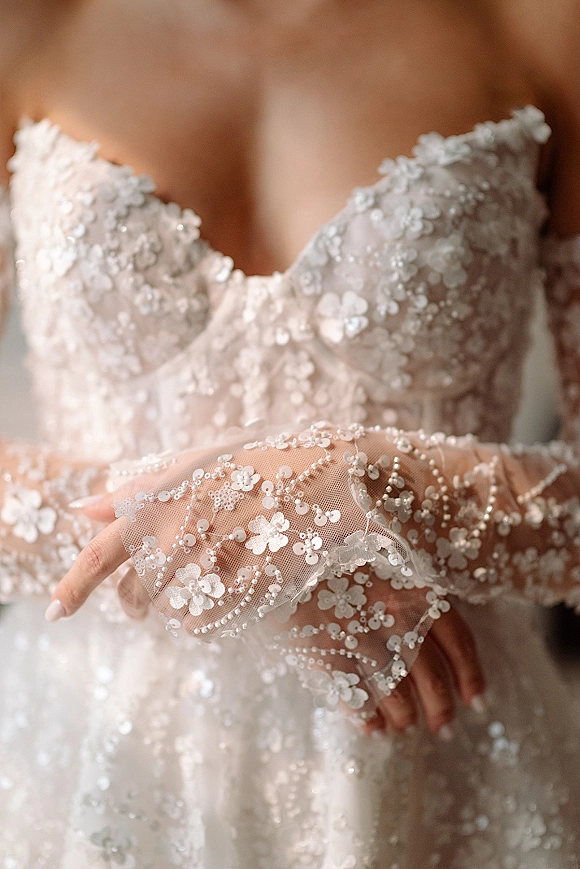 Bridal dress details on a lace wedding dress with strapless floral appliqué bodice, pearl and sequin beading, and sheer tulle gloves indoors