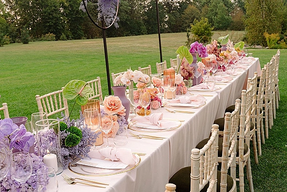 Reception tablescape on a long banquet wedding table with pastel orchids and roses, gold-rim plates, and glass vases on an outdoor lawn