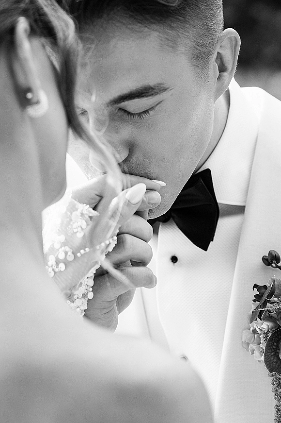 Wedding couple portrait of groom kissing bride’s hand, his white tuxedo and boutonniere beside her lace sleeve and veil in greenery