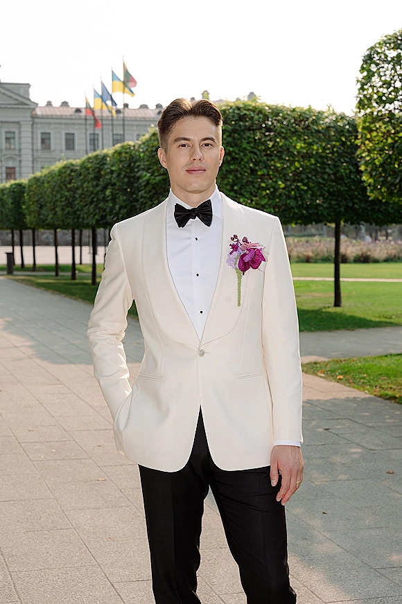 Groom portrait in a white tuxedo jacket with black bow tie and orchid boutonniere, hands in pockets on a garden walkway by hedges