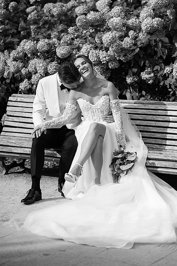 Couple portrait of groom kissing bride’s cheek as she laughs on a wooden bench, veil and bouquet visible beside hydrangea bushes
