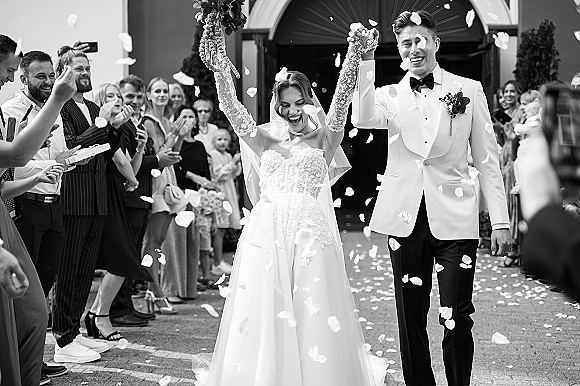 Wedding recessional as newlyweds walking out hand in hand, bride in lace sleeves and veil with groom in tuxedo, confetti petals flying at arched entrance