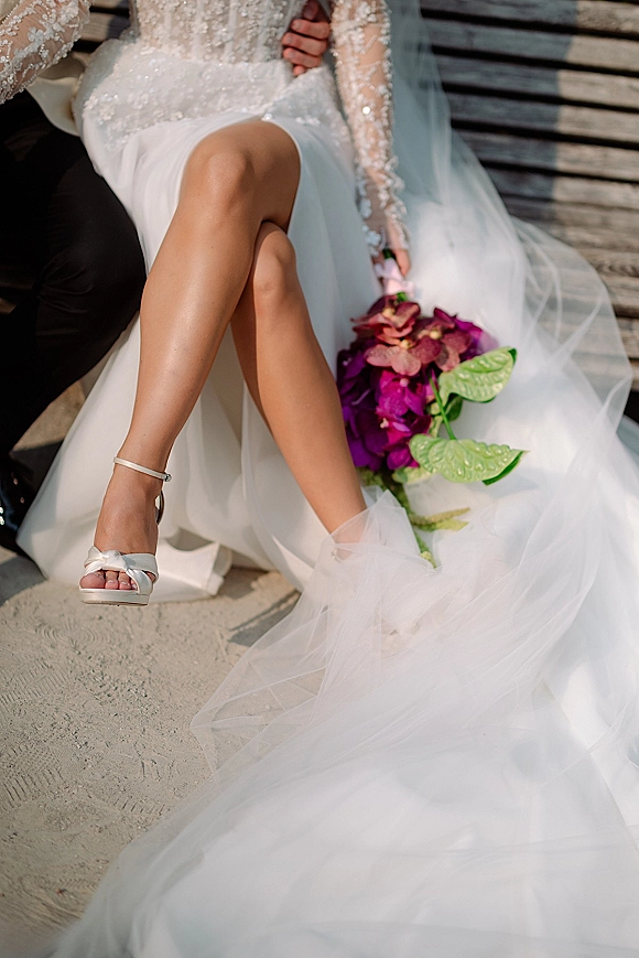 Wedding couple portrait of bride sitting on groom’s lap, veil draped, holding a purple bouquet on a wood bench over sandy ground