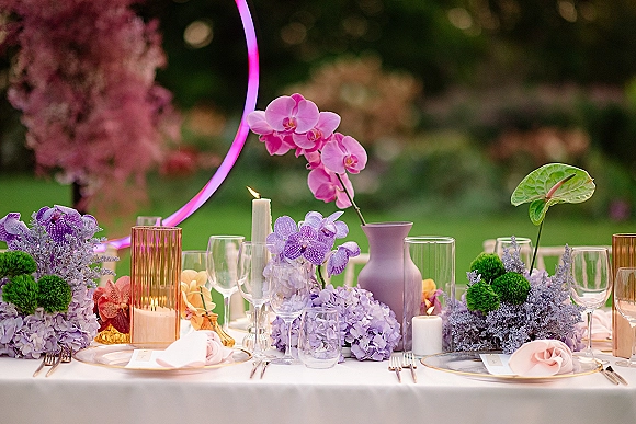 Reception tablescape with orchids and hydrangeas, taper and pillar candles, pastel florals, and place settings on a lawn with neon circle backdrop