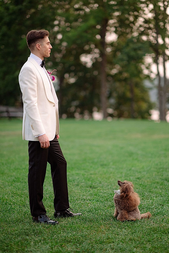 Groom portrait in a white tuxedo standing with his dog on a leash, black bow tie and boutonniere, on a grassy park lawn with trees