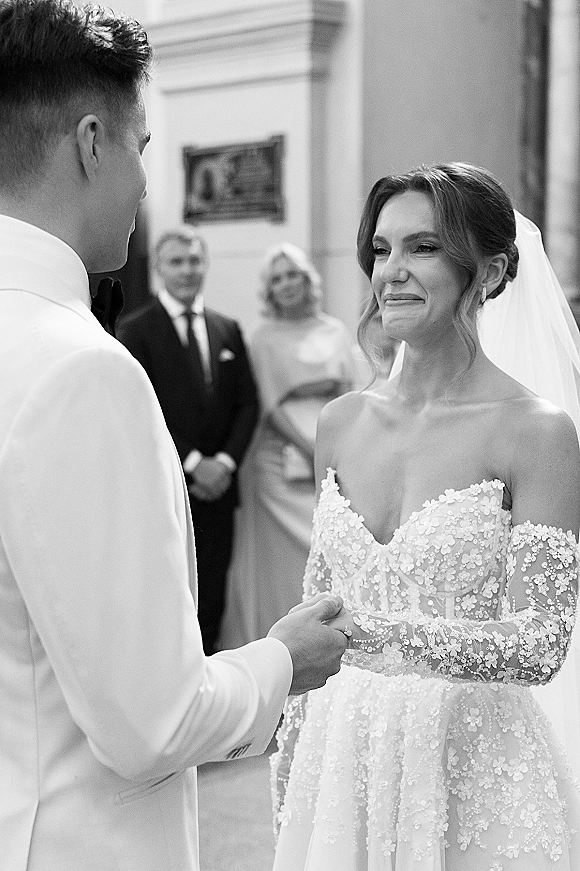 Wedding vows as couple exchanging vows, bride smiling through tears in off-the-shoulder lace and veil, groom in bow tie with guests behind