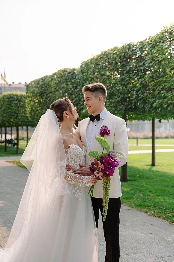 Couple portrait of bride and groom embracing, bride gazing up at him in a park walkway, long veil, lace dress, purple orchid bouquet