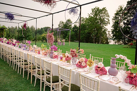 Reception tablescape featuring an outdoor wedding reception table with orchids and hydrangeas, pink napkins, gold flatware under hanging hoops.