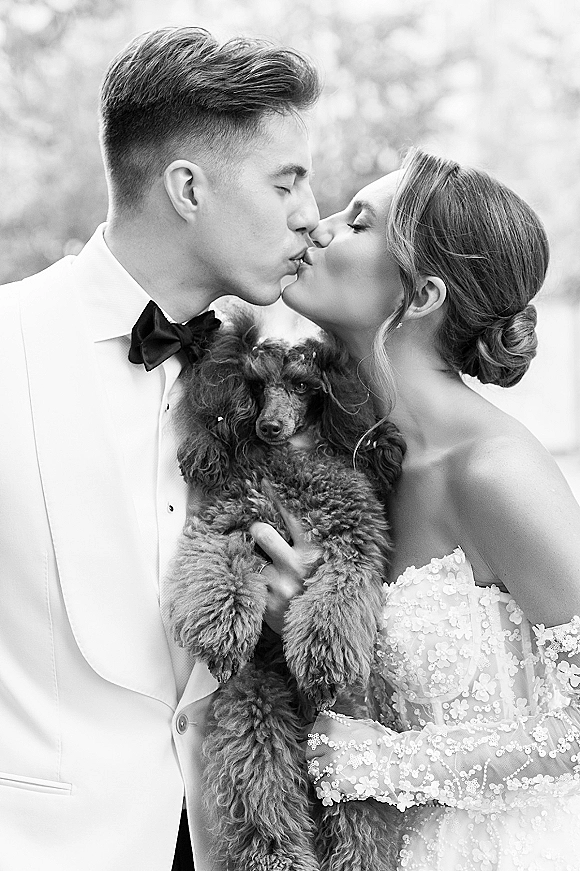 Wedding kiss portrait of bride and groom kissing while holding their dog, lace sleeves and bow tie framed by outdoor greenery and trees