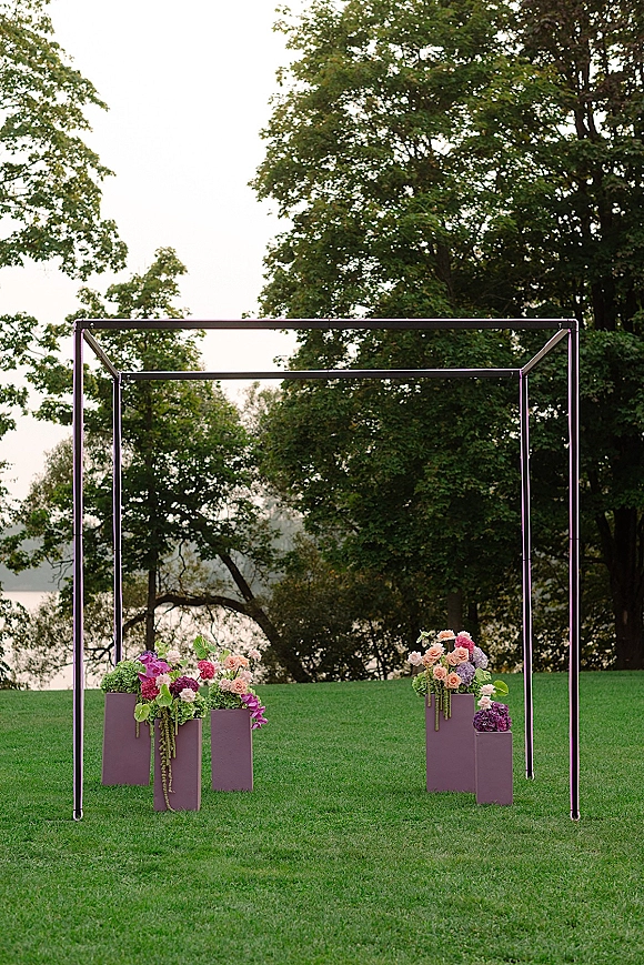 Wedding ceremony backdrop with a square wedding arch, lush roses and orchids on plinths, plus hanging greenery on a lakeside lawn