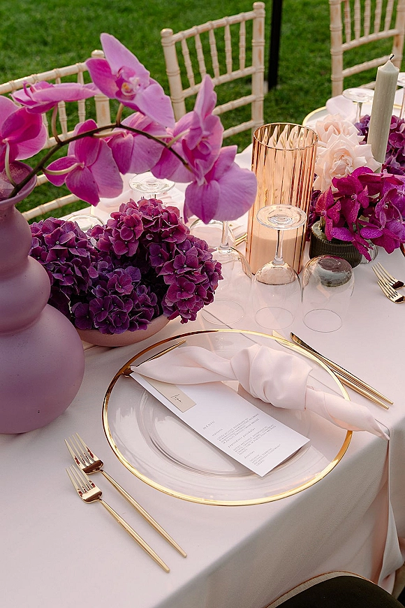 Reception tablescape with purple wedding tablescape orchids, hydrangeas and pink roses, white taper candle, gold charger and flatware on lawn setting