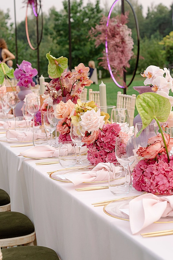 Reception tablescape with wedding table centerpiece of orchids and hydrangea, gold chargers, blush napkins, taper candles on an outdoor lawn under trees