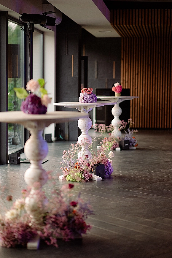 Cocktail hour decor with wedding cocktail tables topped with bud vases and floral centerpieces, set by floor-to-ceiling windows and uplights