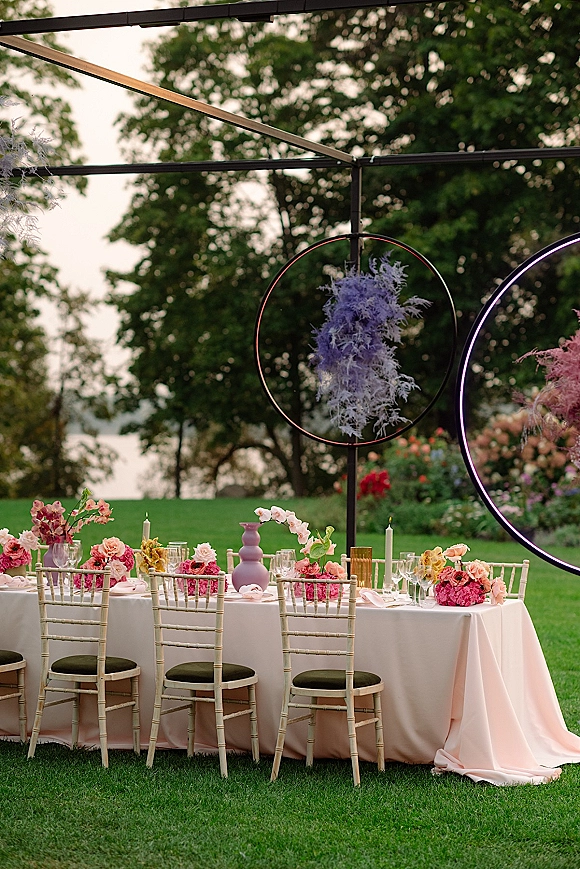 Reception tablescape with blush linen, pastel vases, orchid stems, taper candles and glassware, set on a green lawn with neon circle backdrop
