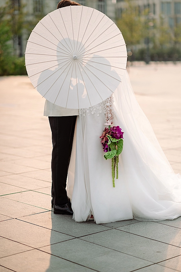 Wedding couple portrait with bride and groom holding a white paper parasol, hugging in an outdoor plaza; veil trails behind purple bouquet.