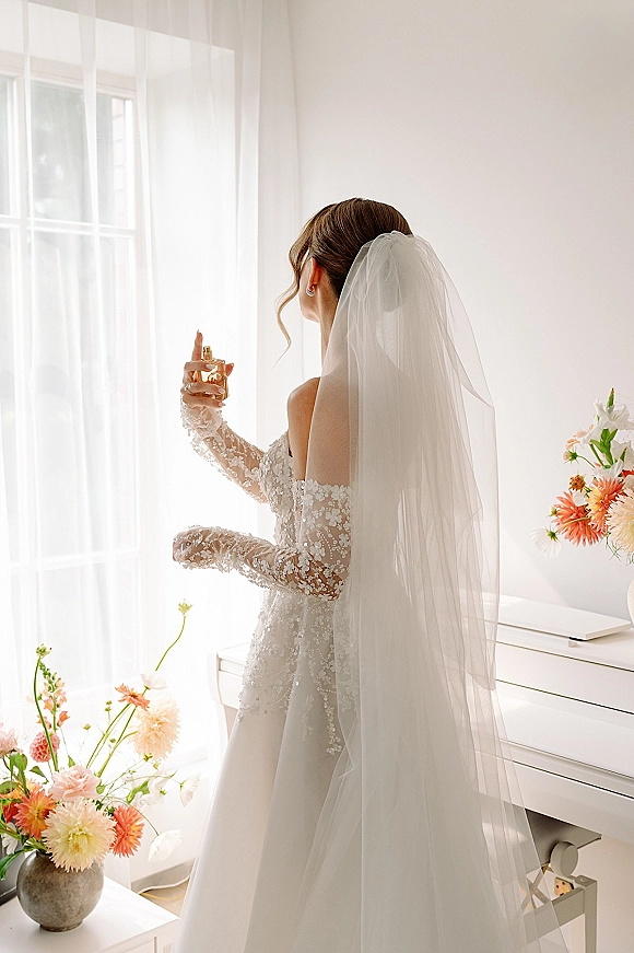 Bridal getting ready, bride spraying perfume in an off-the-shoulder lace wedding dress and long veil by a bright window, flowers on piano