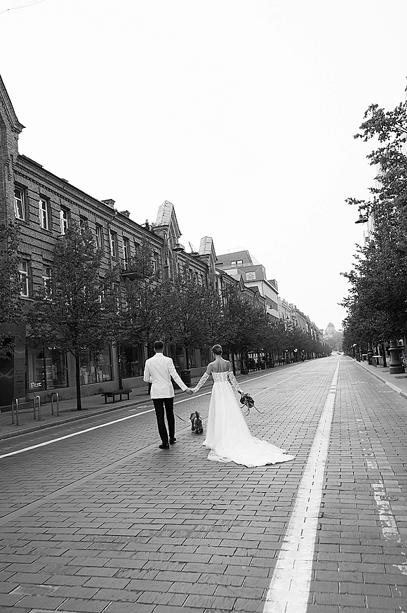Couple portrait in a black and white wedding photo, bride and groom walking away hand in hand with dog on leash on empty city street