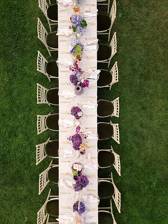 Reception tablescape with an outdoor reception table set with purple and blue florals, gold-rim chargers, menus, and glassware on a lawn