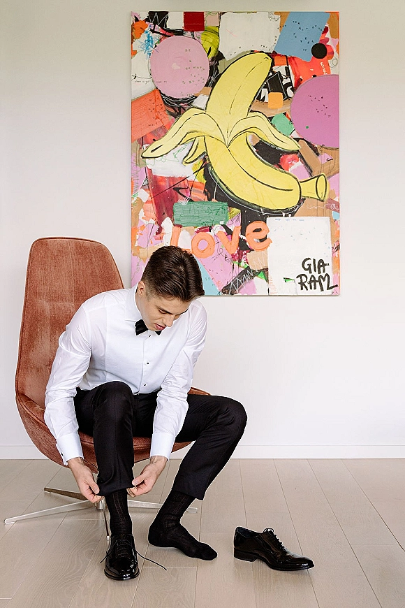 Groom getting ready tying shoes, seated in a brown chair with black patent leather dress shoes and bow tie by abstract painting