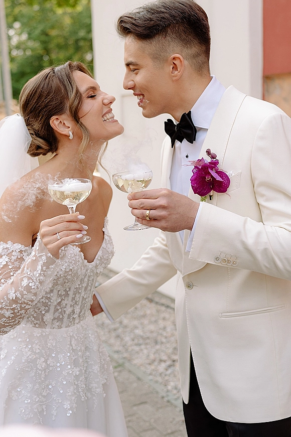 Wedding couple portrait with champagne toast, bride and groom laughing while holding coupe glasses in a green courtyard by a white wall