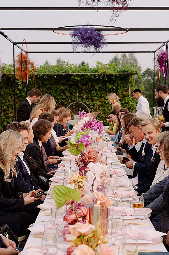 Reception tablescape with a long banquet table wedding setup, floral centerpieces and candles under a metal frame in a garden hedge backdrop