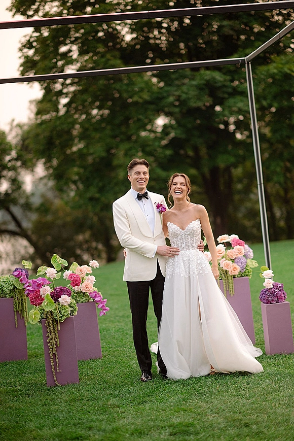 Couple portrait of bride in strapless slit wedding dress and groom in white dinner jacket laughing beneath floral pedestals on a lawn