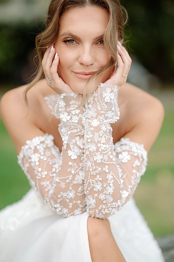Bridal portrait of a bride with natural bridal makeup and a loose updo, wearing floral lace gloves and a strapless dress in a blurred garden backdrop