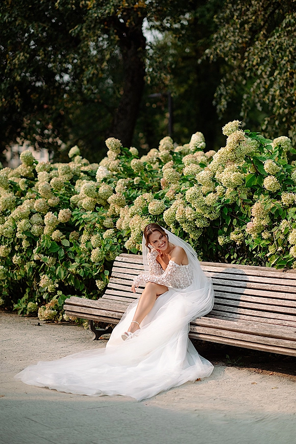 Bridal portrait of a bride sitting on a wooden bench in a park, wearing a long veil and lace-sleeve tulle wedding dress amid flowering bushes