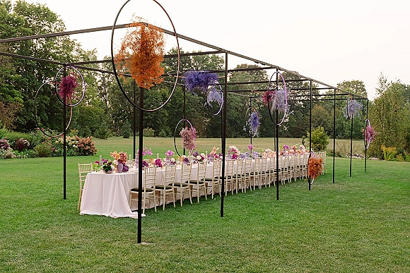 Reception tablescape outdoor banquet table with white tablecloth, chiavari chairs, floral centerpieces, candles, purple glassware under hanging hoops on a lawn