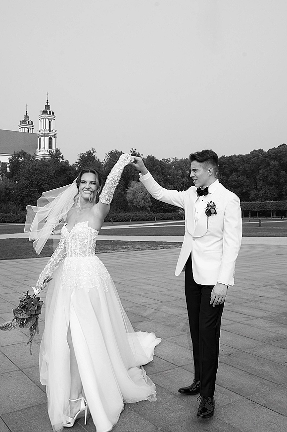 Couple portrait, black and white wedding portrait of groom spinning laughing bride in veil and lace gloves on an outdoor plaza by a church