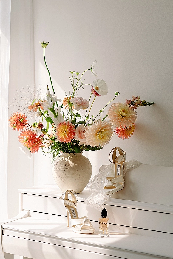 Bridal accessories with white strappy heels, hair bow, and perfume bottle beside colorful flowers on a sunlit white dresser
